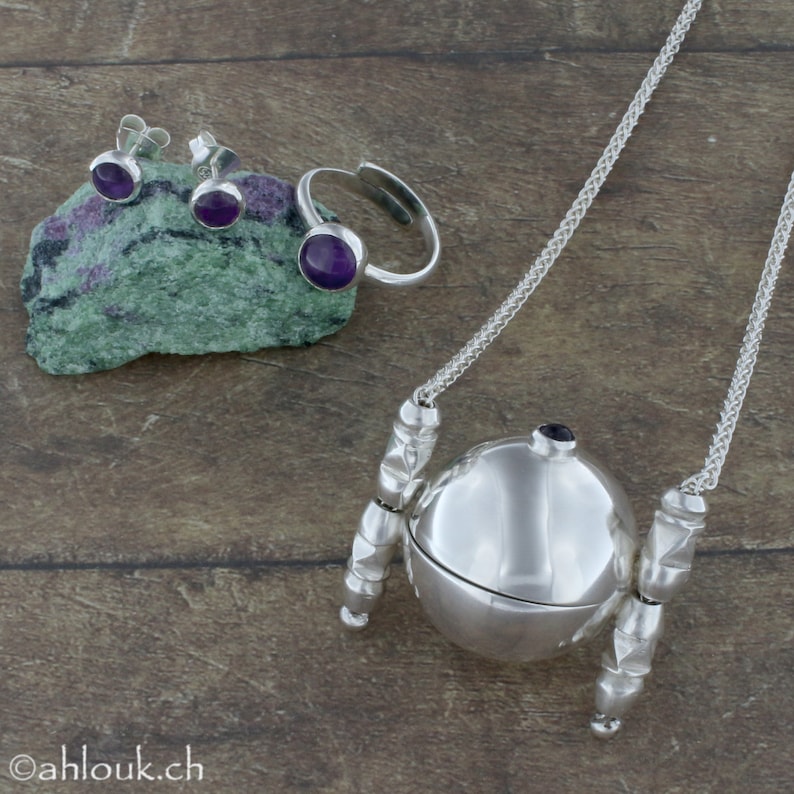 May include: A set of silver jewelry featuring amethyst gemstones. The set includes a pair of earrings, a ring, and a pendant necklace with a round, hinged locket. The locket is decorated with a silver chain and a silver ball.