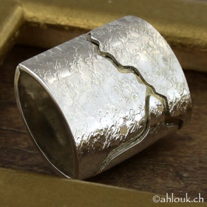 May include: A wide, textured silver ring with a unique split design. The ring has a rough, hammered appearance and a crack-like detail that runs across the band. The ring is displayed on a wooden surface.
