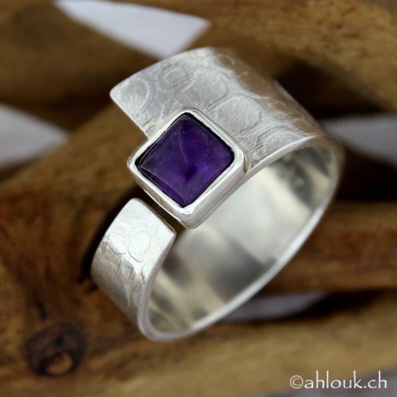 May include: A silver ring with a square amethyst gemstone set in a textured band. The ring is open and adjustable.