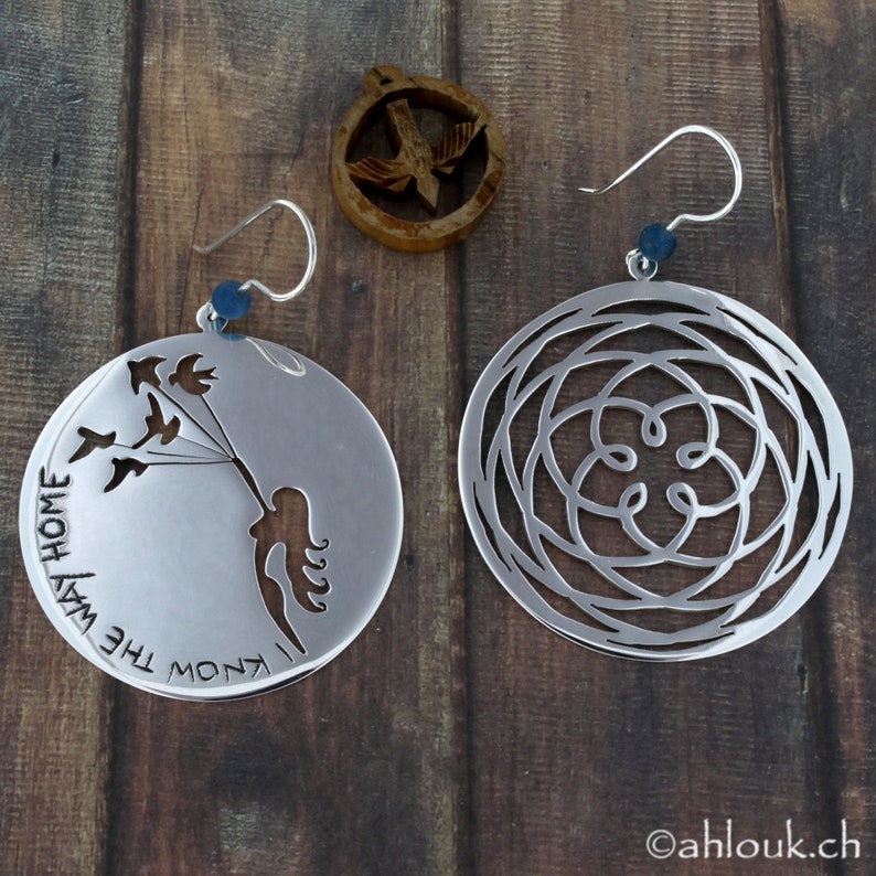 May include: A pair of silver earrings. One earring features a cutout design of a woman holding a string of birds with the text "Know the way home". The other earring features a circular design with a repeating pattern.