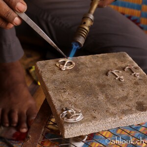 May include: A person uses a blowtorch to solder silver anchor charms on a gray stone surface. The charms are arranged in a row, with one being soldered.