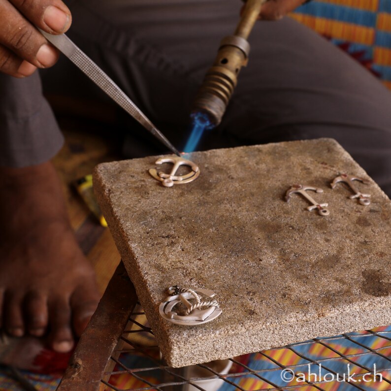 May include: A person uses a blowtorch to solder silver anchor charms on a gray stone surface. The charms are arranged in a row, with one being soldered.