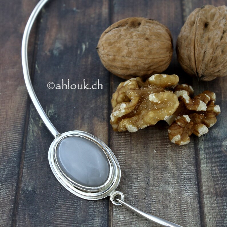 Peut inclure: Un collier en argent avec un grand pendentif ovale en pierre grise. Le collier est sur une surface en bois avec des noix.