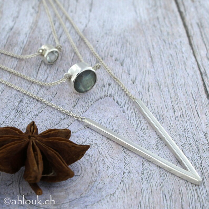 May include: A silver necklace with a V-shaped pendant and two smaller round pendants. The necklace is on a wooden surface with a star anise.