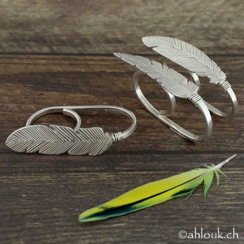 May include: Two silver feather rings, one with a single feather and the other with three stacked feathers. The rings are on a wooden surface with a yellow and green feather.