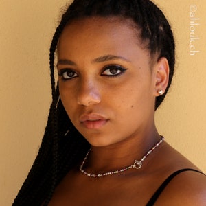 May include: A young woman with long black braids wears a silver necklace with small red, white, and clear beads. She is wearing a black tank top and has a neutral expression.