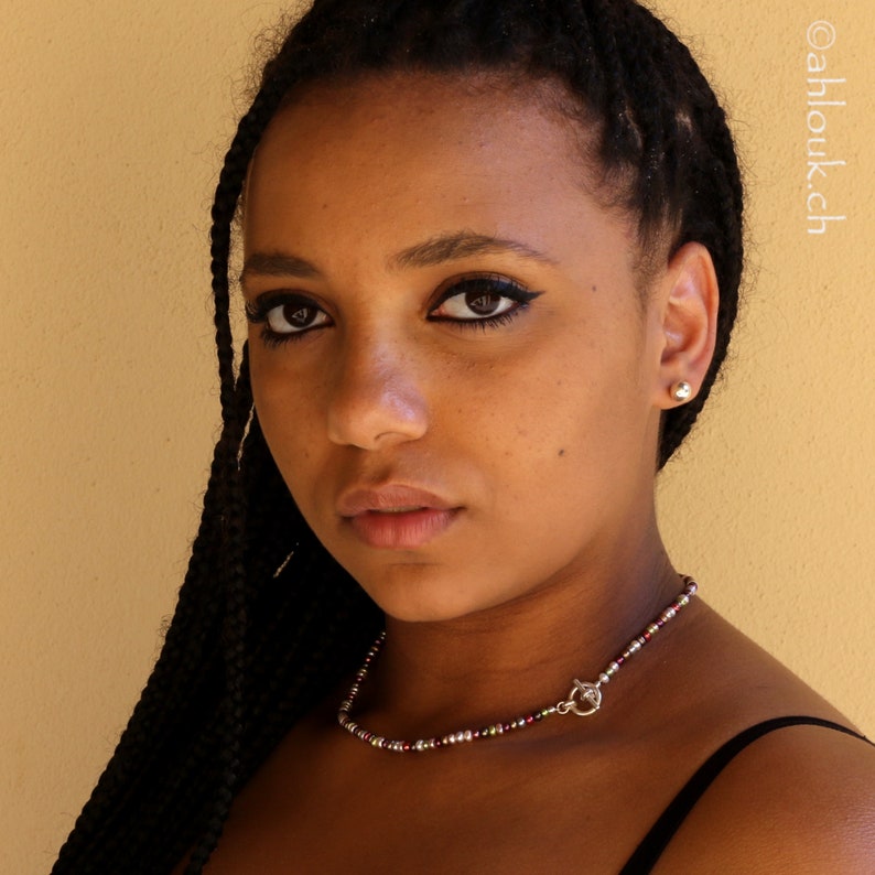 May include: A young woman with long black braids wears a silver necklace with small red, white, and clear beads. She is wearing a black tank top and has a neutral expression.