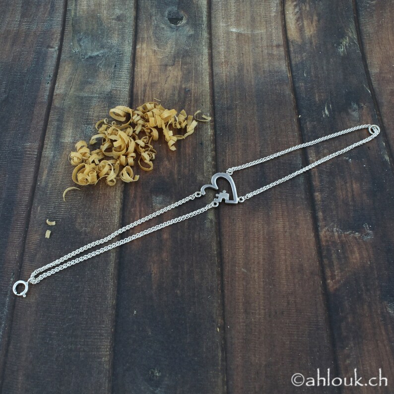 May include: A silver chain necklace with a heart-shaped pendant featuring a puzzle piece design. The necklace is on a wooden surface.