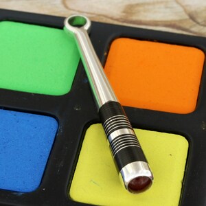 May include: A silver and black metal tool with a red gemstone at the end, lying on a black tray with colorful squares.