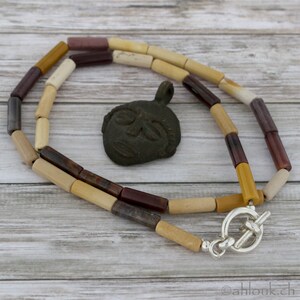 May include: A necklace made of beige, brown, and red cylindrical beads with a silver toggle clasp. The necklace is lying on a wooden surface with a small, dark brown, carved face pendant.