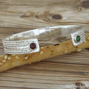 Puede incluir: Pulsera de plata con un dise&ntilde;o trenzado y dos acentos de piedras preciosas. Una piedra preciosa es roja y la otra es verde.