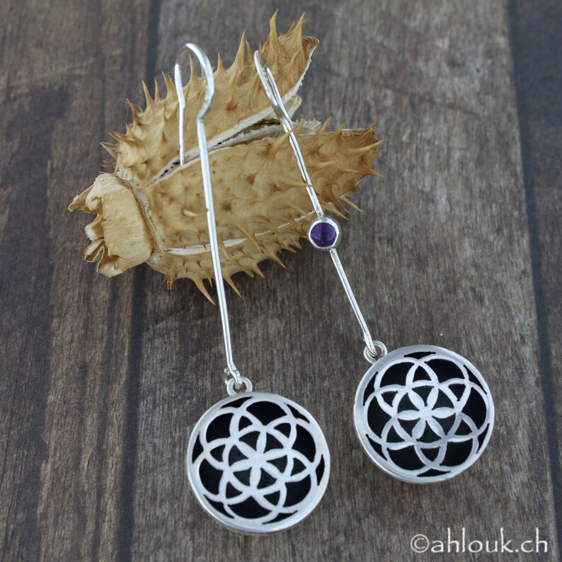 May include: A pair of silver earrings with a black and silver geometric design and a small purple gemstone. The earrings are hanging from a silver hook.