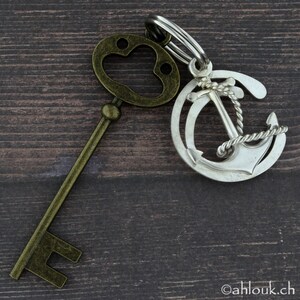 May include: A silver anchor charm with a rope design attached to a gold key with a paw print on the top. The charm and key are on a wooden surface.