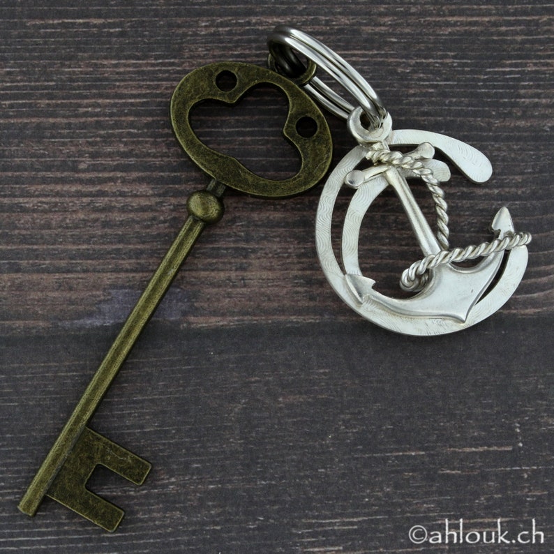 May include: A silver anchor charm with a rope design attached to a gold key with a paw print on the top. The charm and key are on a wooden surface.