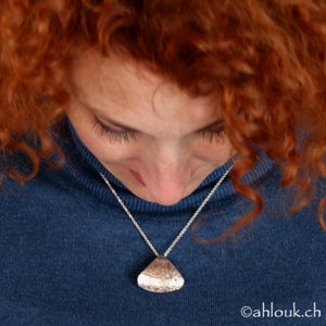 May include: A silver necklace with a unique, textured pendant. The pendant is a light, metallic color and hangs from a delicate silver chain. The person wearing the necklace is wearing a blue turtleneck sweater.