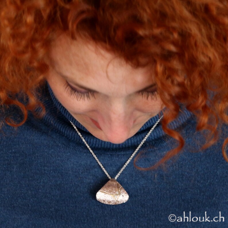 May include: A silver necklace with a unique, textured pendant. The pendant is a light, metallic color and hangs from a delicate silver chain. The person wearing the necklace is wearing a blue turtleneck sweater.