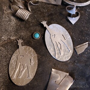 May include: Close-up of jewelry-making tools and components. Two oval-shaped silver pendants with giraffe silhouettes and heart cutouts. A small turquoise stone is set in a silver bezel. Various metal scraps and coils are scattered around.