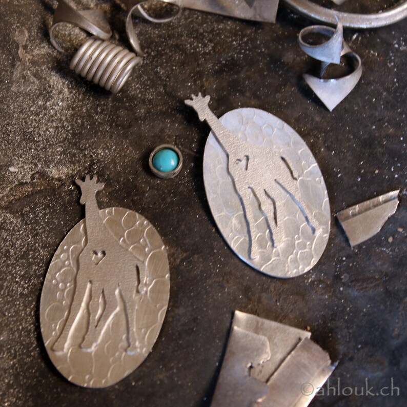 May include: Close-up of jewelry-making tools and components. Two oval-shaped silver pendants with giraffe silhouettes and heart cutouts. A small turquoise stone is set in a silver bezel. Various metal scraps and coils are scattered around.