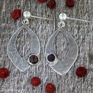 May include: A pair of silver earrings with a textured, leaf-like design. Each earring features a red gemstone in the center.
