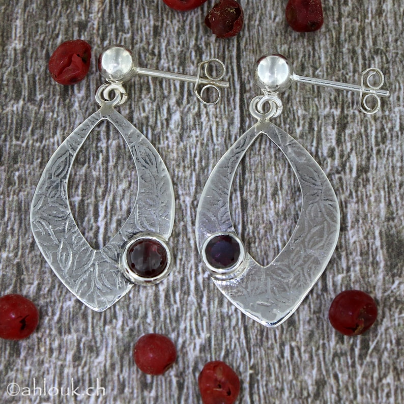 May include: A pair of silver earrings with a textured, leaf-like design. Each earring features a red gemstone in the center.