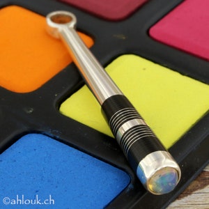 May include: A silver and black pen with a clear opal gemstone at the end. The pen is lying on a colorful paint palette.