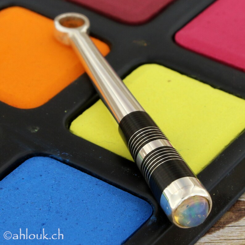 May include: A silver and black pen with a clear opal gemstone at the end. The pen is lying on a colorful paint palette.