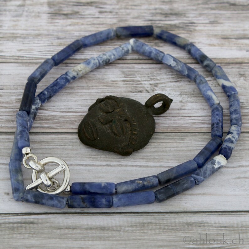 May include: A blue and white beaded necklace with a silver toggle clasp. The necklace is lying on a wooden surface next to a small, dark brown pendant in the shape of a face.