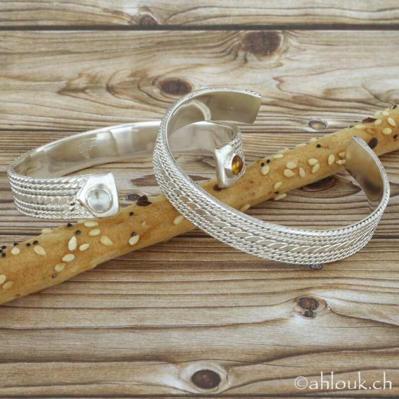 Puede incluir: Dos pulseras de plata con dise&ntilde;os intrincados y peque&ntilde;as piedras preciosas. Las pulseras est&aacute;n apoyadas sobre una superficie de madera con una barrita de pan.