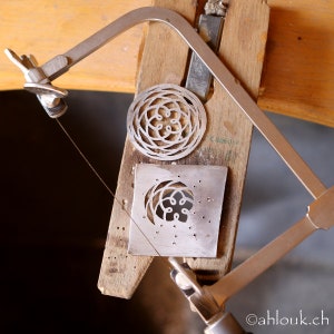 May include: A close-up of a metalworking project in progress. A silver metal piece with a circular design is being cut with a hand-held saw. The saw is mounted on a wooden frame.