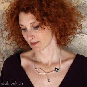 May include: A woman with red curly hair wears a silver necklace with a green leaf design. The necklace has small, round, yellow gemstones.