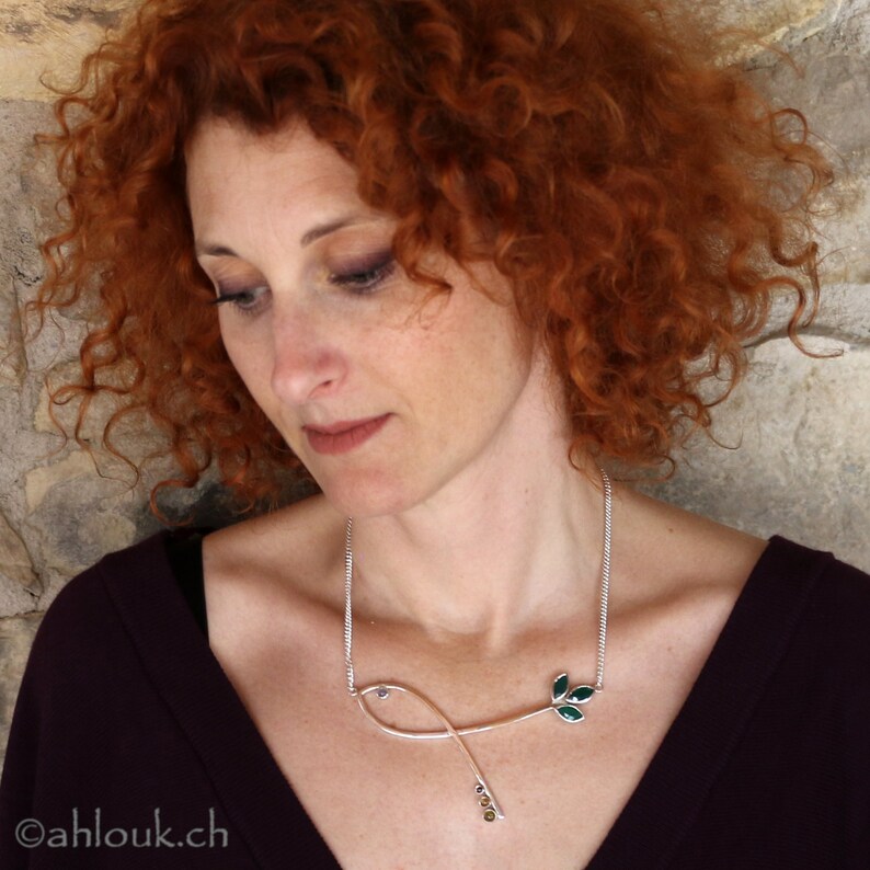 May include: A woman with red curly hair wears a silver necklace with a green leaf design. The necklace has small, round, yellow gemstones.