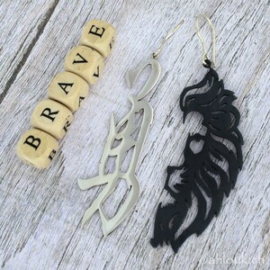 May include: A pair of earrings with a silver and black design. The silver earring is shaped like the word "brave" in a stylized font. The black earring is shaped like a stylized lion's mane.