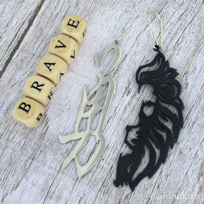 May include: A pair of earrings with a silver and black design. The silver earring is shaped like the word "brave" in a stylized font. The black earring is shaped like a stylized lion's mane.