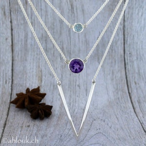 May include: A silver necklace with three chains and two pendants. The top pendant is a small light blue gemstone. The bottom pendant is a larger, round, purple amethyst gemstone. The chains are connected to a V-shaped silver pendant.