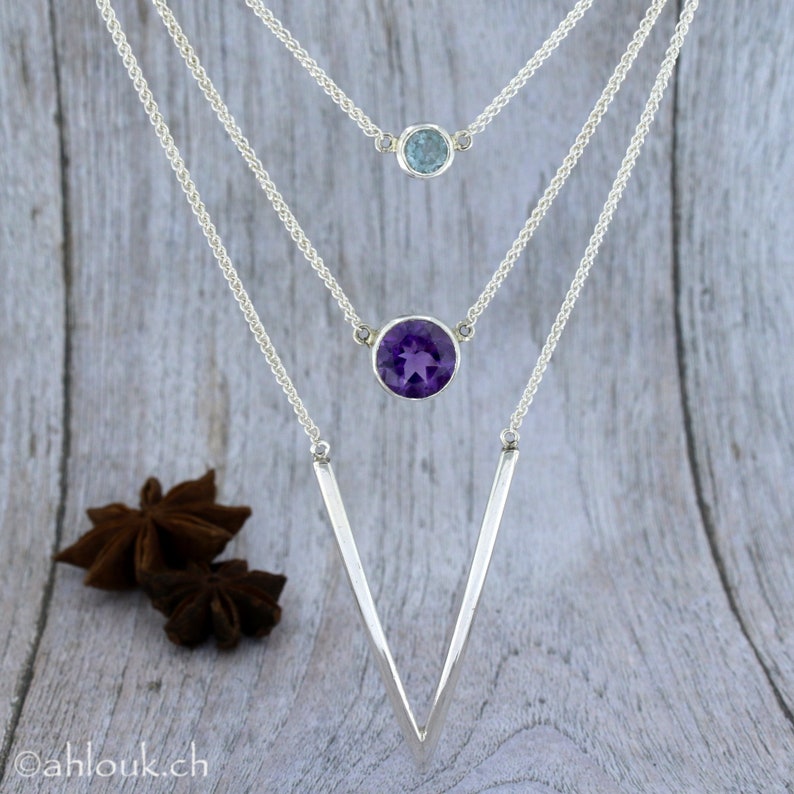 May include: A silver necklace with three chains and two pendants. The top pendant is a small light blue gemstone. The bottom pendant is a larger, round, purple amethyst gemstone. The chains are connected to a V-shaped silver pendant.