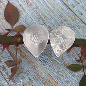 May include: Two silver guitar picks with a rose design. One pick has a single rose in the center, while the other pick has a repeating pattern of roses.