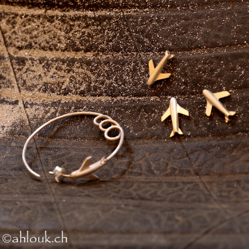 May include: A silver bracelet with three connected rings and a silver airplane charm. Three additional silver airplane charms are scattered on a textured brown surface.