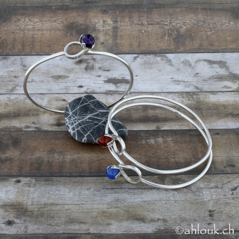 May include: Two silver bangle bracelets with gemstone accents. The top bracelet has a purple gemstone, the bottom bracelet has a red gemstone and a blue gemstone.