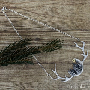 May include: A silver necklace with a pendant featuring a stylized antler design with a crescent moon and a cluster of trees inside. The pendant is textured and has a rustic look.