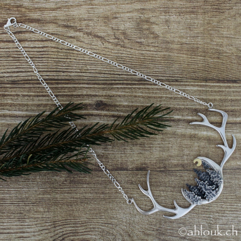 May include: A silver necklace with a pendant featuring a stylized antler design with a crescent moon and a cluster of trees inside. The pendant is textured and has a rustic look.