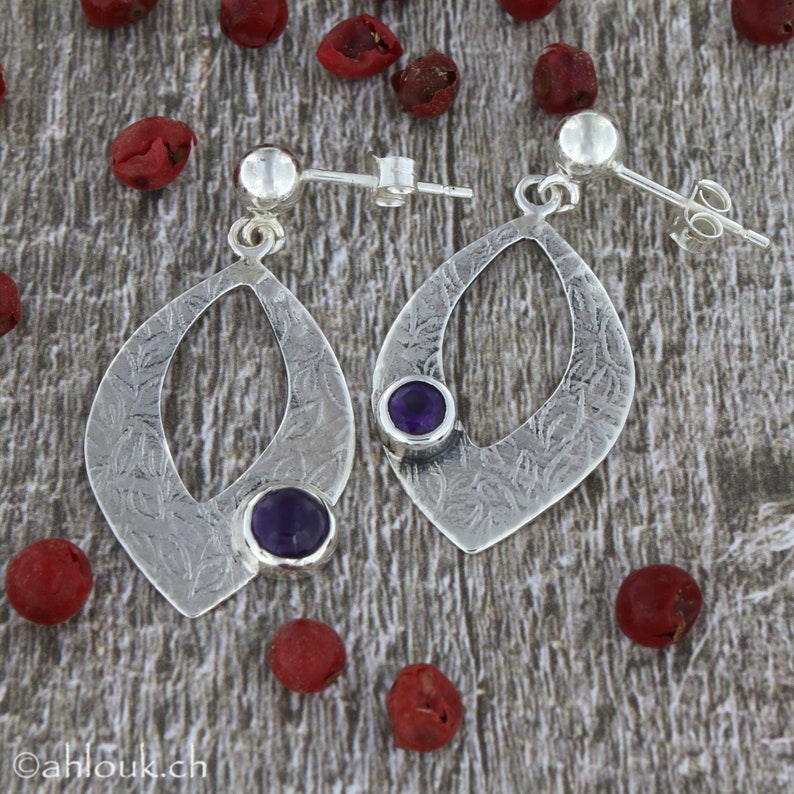 May include: A pair of silver earrings with a textured, leaf-shaped design. Each earring features a small, round, purple amethyst gemstone.
