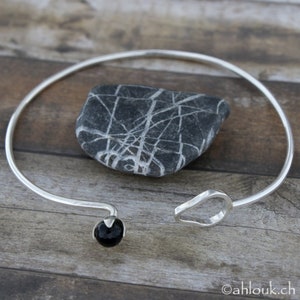 May include: A silver bangle bracelet with a black gemstone bead and a curved, open design. The bracelet is on a wooden surface.