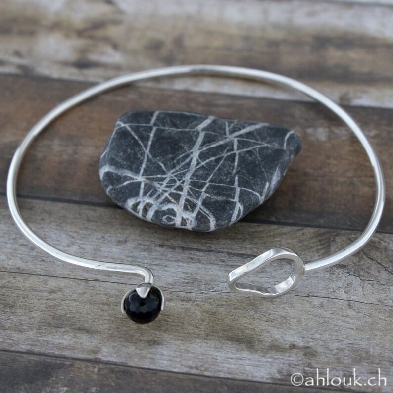 May include: A silver bangle bracelet with a black gemstone bead and a curved, open design. The bracelet is on a wooden surface.