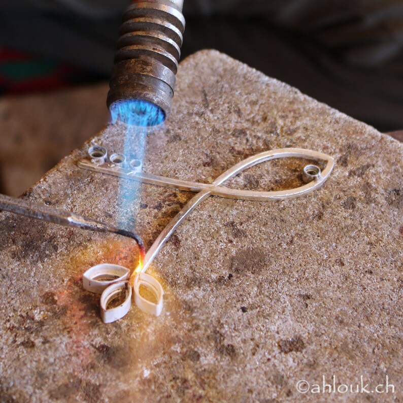 May include: A silver fish-shaped pendant is being soldered with a torch. The pendant has a small loop at the top and a small circle in the middle. The torch is held by a hand, but the hand is not visible in the image.
