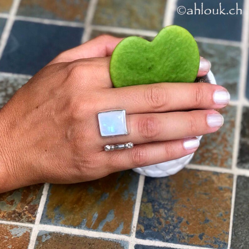 May include: A silver ring featuring a large, rectangular, iridescent moonstone set in a bezel setting. The ring is worn on the index finger of a hand.