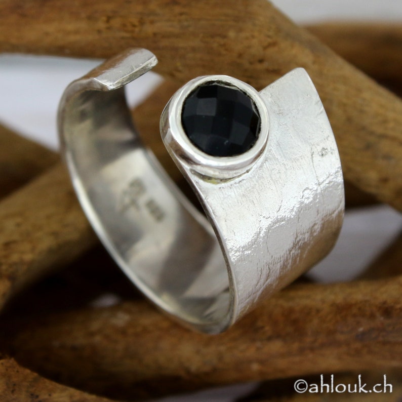 May include: A silver ring with a black gemstone set in a textured band. The ring is open on one side and has a hammered finish.