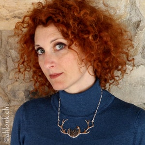 May include: A woman with curly red hair wearing a blue turtleneck sweater and a silver necklace with a deer antler pendant.