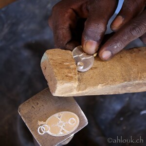 May include: A silversmith works on a silver pendant with an oval shape and intricate geometric designs. The pendant is held in place with a wooden tool. The artisan's hands are visible, demonstrating the craftsmanship involved in jewelry making.