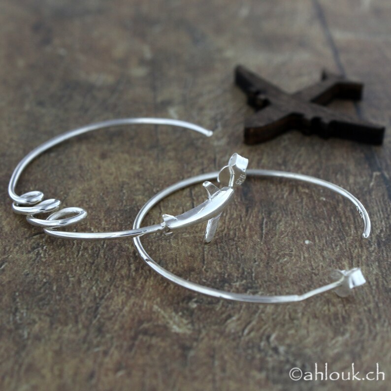 May include: A pair of silver hoop earrings, one with a small airplane charm. The earrings are on a wooden surface.