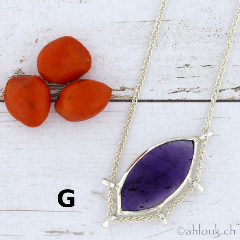 Puede incluir: Un collar de plata con un colgante de piedra preciosa p&uacute;rpura. El colgante tiene forma de l&aacute;grima y est&aacute; engastado en plata. El collar est&aacute; sobre una superficie de madera blanca con tres semillas rojas.